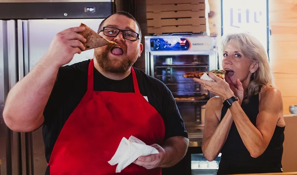 The owners of New York Pizza Depot behind the counter, each eating a slice
